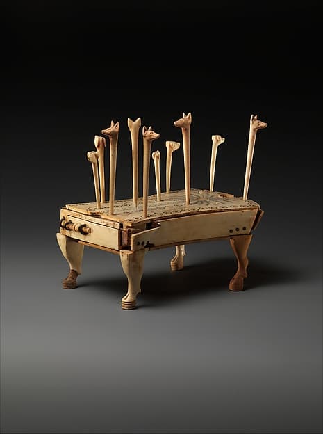 A small game table on carved legs, a palm tree on its shield-shaped top, with ivory pegs carved as hound and jackal heads standing in rows of drilled holes