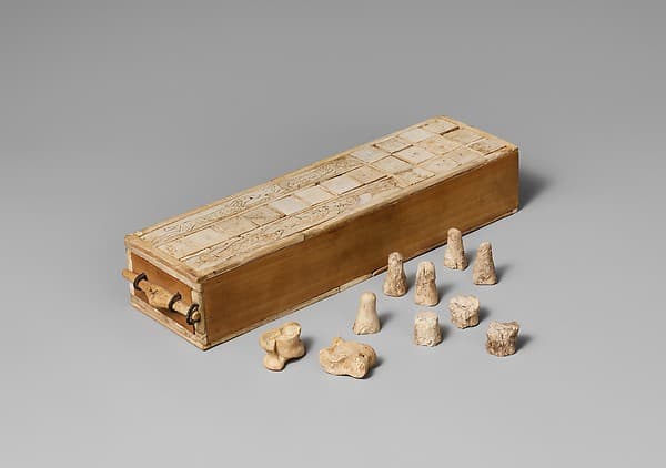 An ancient Egyptian ivory-veneered game box with a small drawer, its top ruled into the thirty squares of senet, gaming pieces beside it